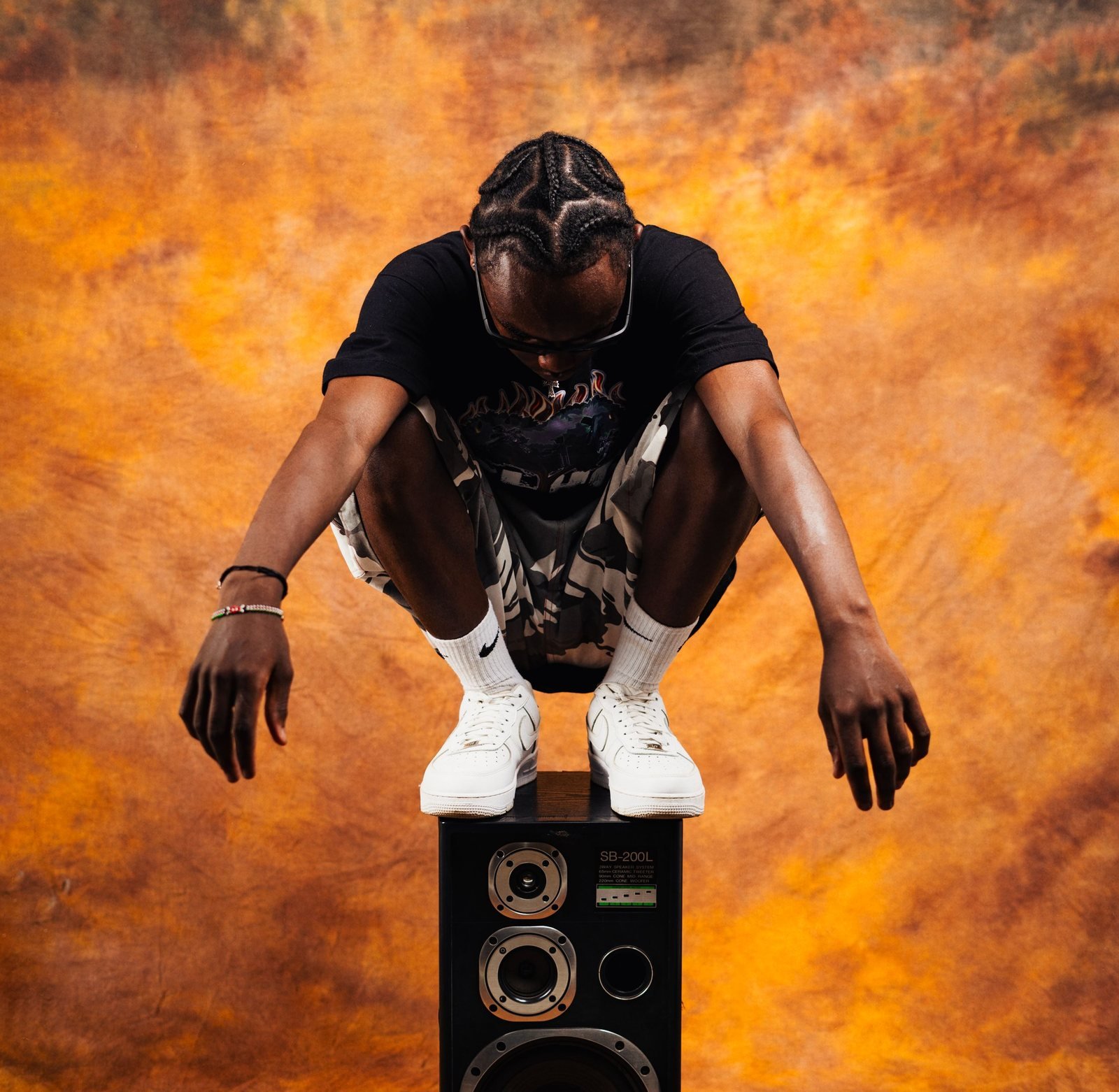 A man crouching on top of a floor-standing loudspeaker in front of a warm backdrop
